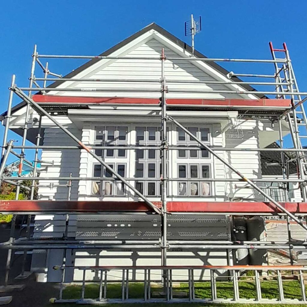 The image is of a white building with a dark roof, set against a clear sky. The house is shown with scaffolding around it, indicating that it is being prepared to paint