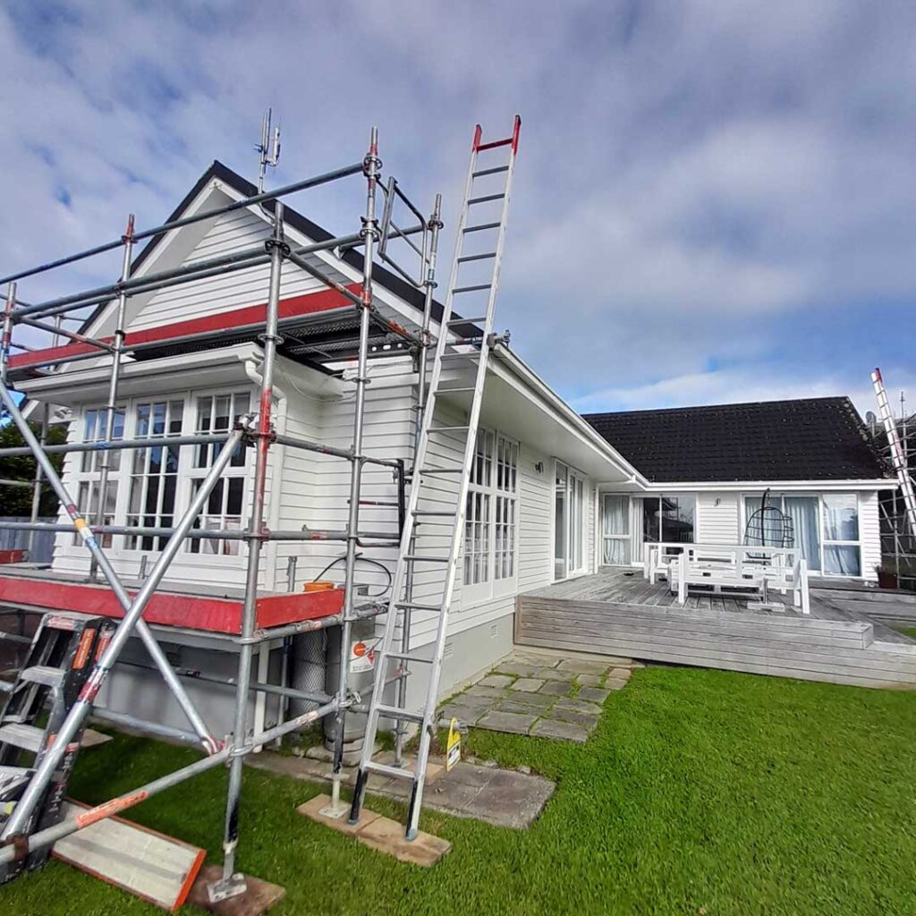 The image depicts a white house. The house is surrounded by a grassy area with a ladder and scaffolding visible, indicating that the house is being prepared for painting.