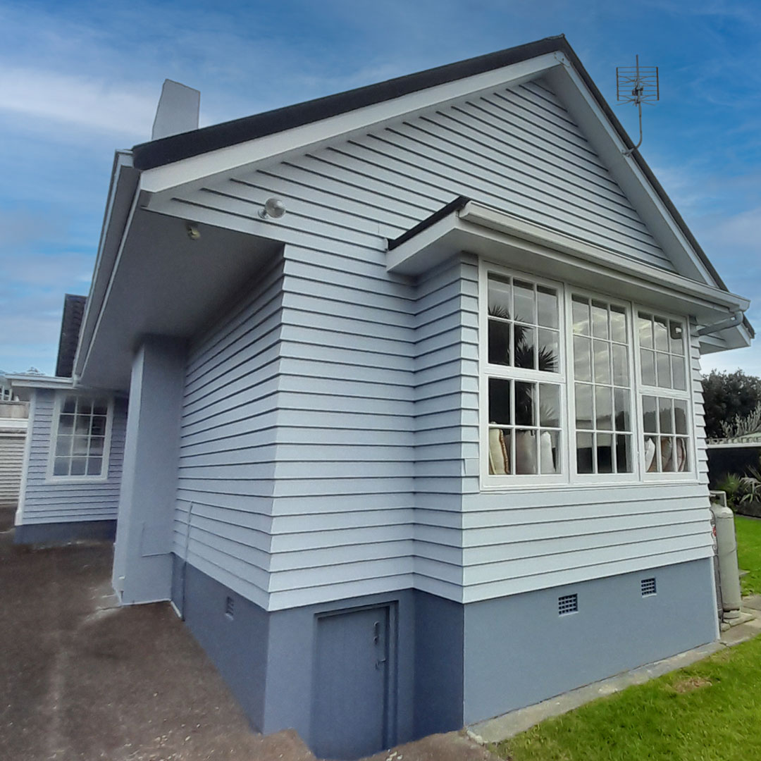 The image shows a white house with a black roof. The house has a porch and is surrounded by grass and a garden shed. The sky is visible in the background, with some clouds. The photo is related to exterior house painting.