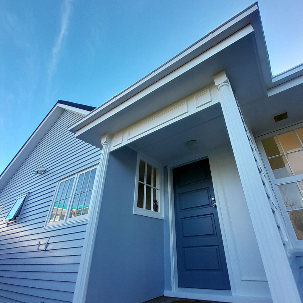 The image is of a white house with a grey door, taken after exterior house painting. It features a clear blue sky in the background.