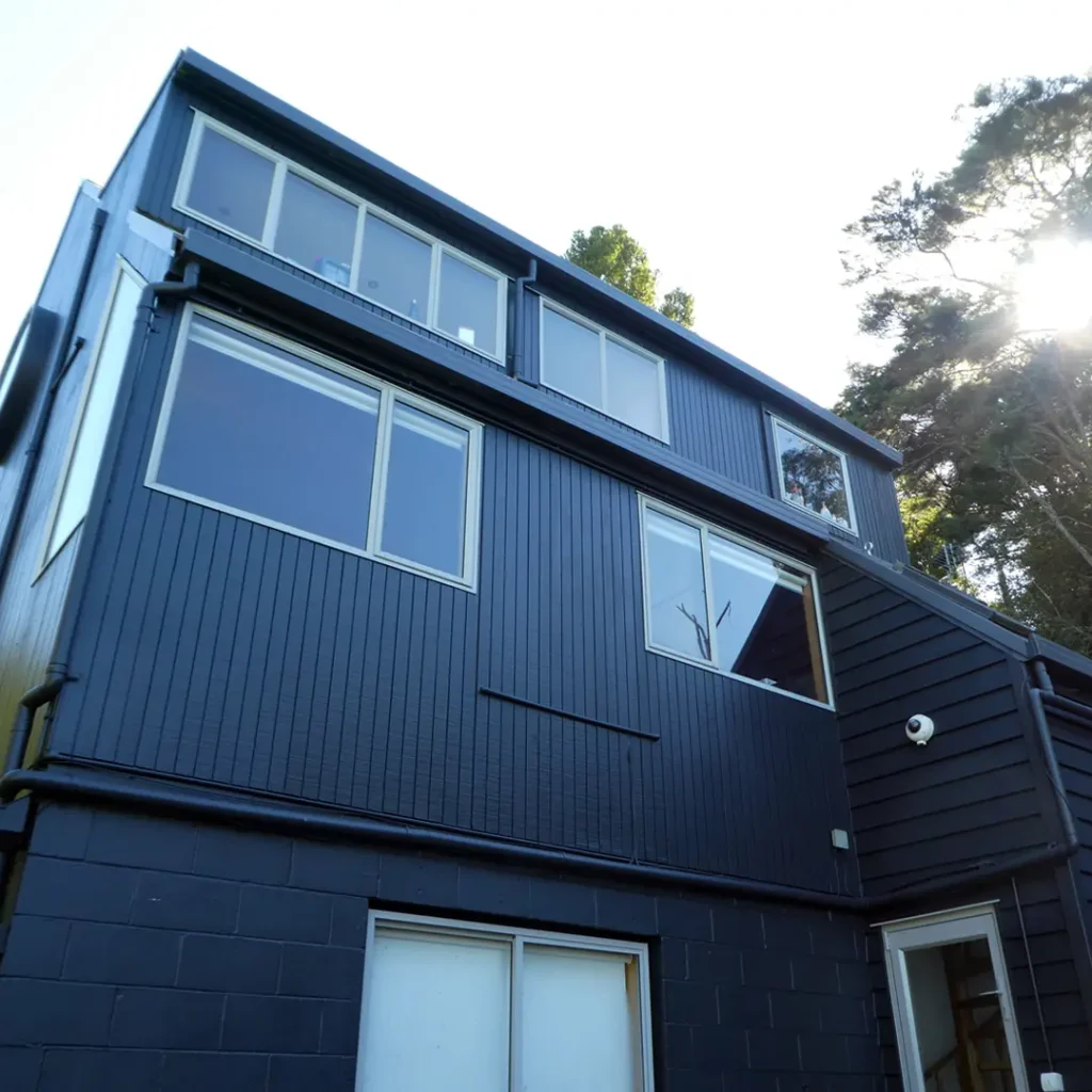 The picture shows a newly painted house. It has a roof, vertical cladding, aluminium windows, and a tree in the distance. Weathershield® ColourGuard™ Rawene. paint gives the house a fresh appearance.