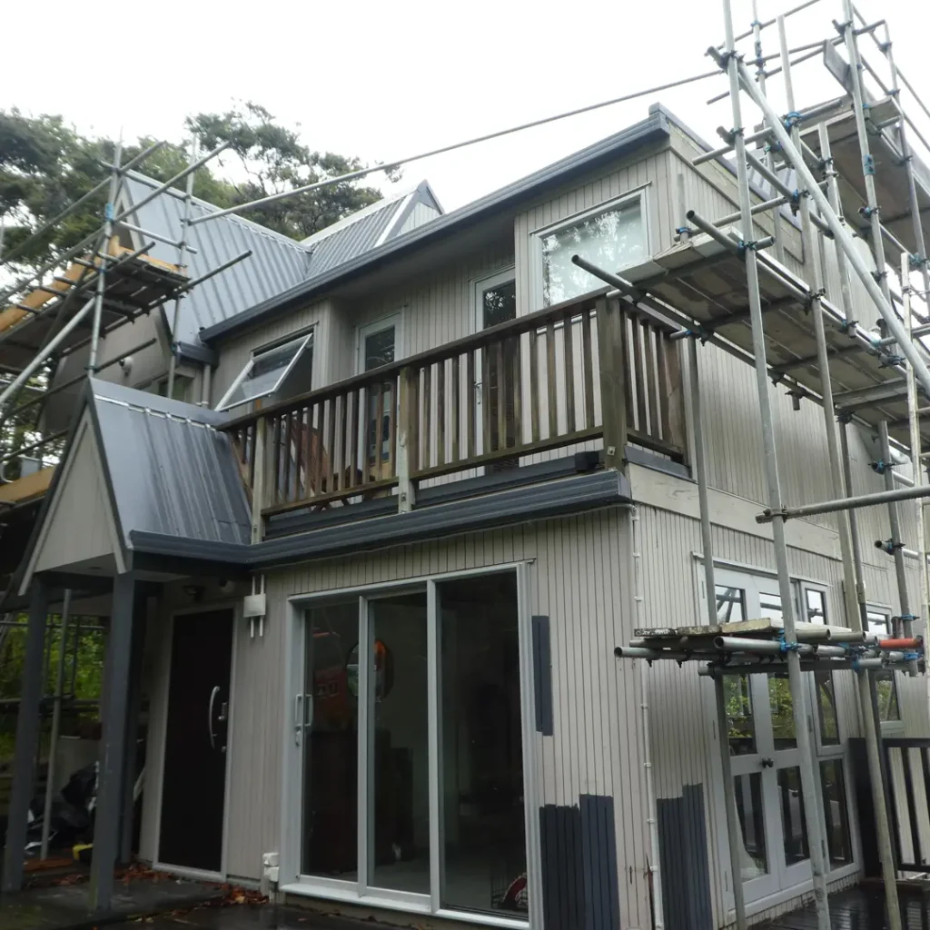 The image depicts a house showing the building process before exterior painting. The scene includes a deck, scaffolding, roof, windows, and vertical cladding, set against a backdrop of trees and the sky.