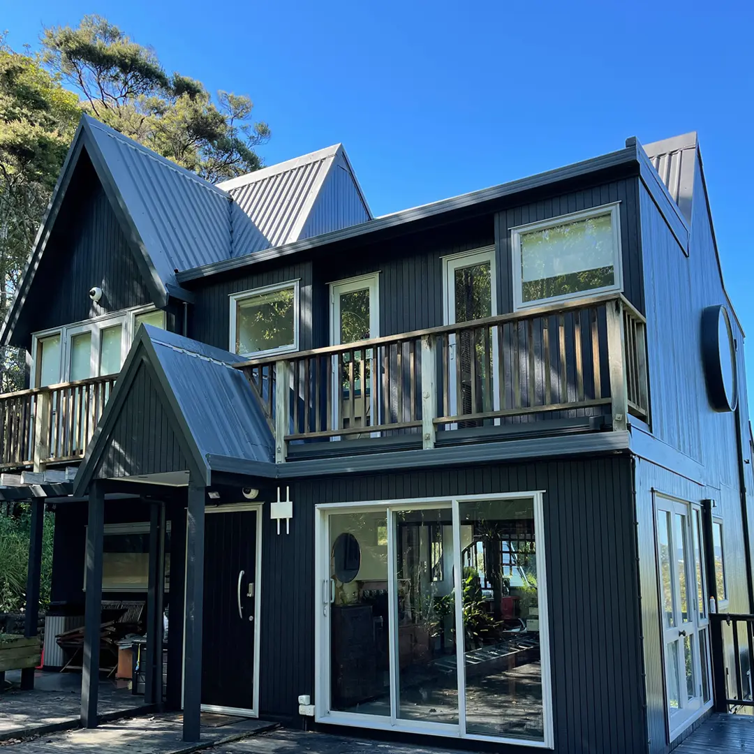 The image is of a house with a deck. It features aluminium windows, vertical cladding, a roof, and a tree in the background. The house looks newly painted with Weathershield® ColourGuard™ Rawene.