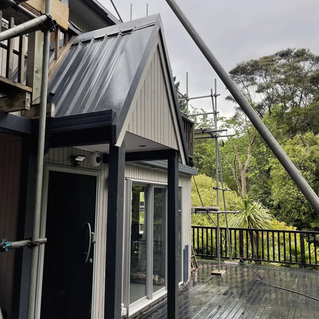 The picture shows a house in the stages leading up to exterior painting. A deck, scaffolding, roof, aluminum windows, and vertical cladding are all visible in the scene, which is framed by trees and the sky.