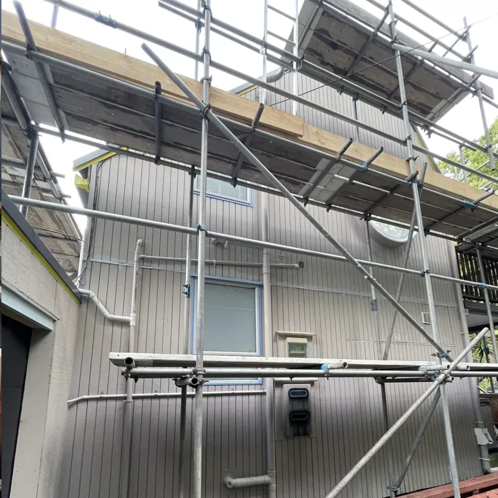 The image depicts a house in preparation for its exterior paint job. The scene is framed by trees and the sky and features a deck, scaffolding, roof, aluminum windows, and vertical cladding.