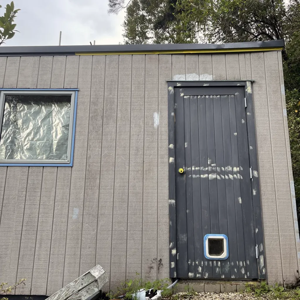 The photo shows a wood building with a black door, situated outdoors. The building appears to be in its original state before exterior painting. Other elements in the image include a tree, windows, a waste container, a plant, and a sign.