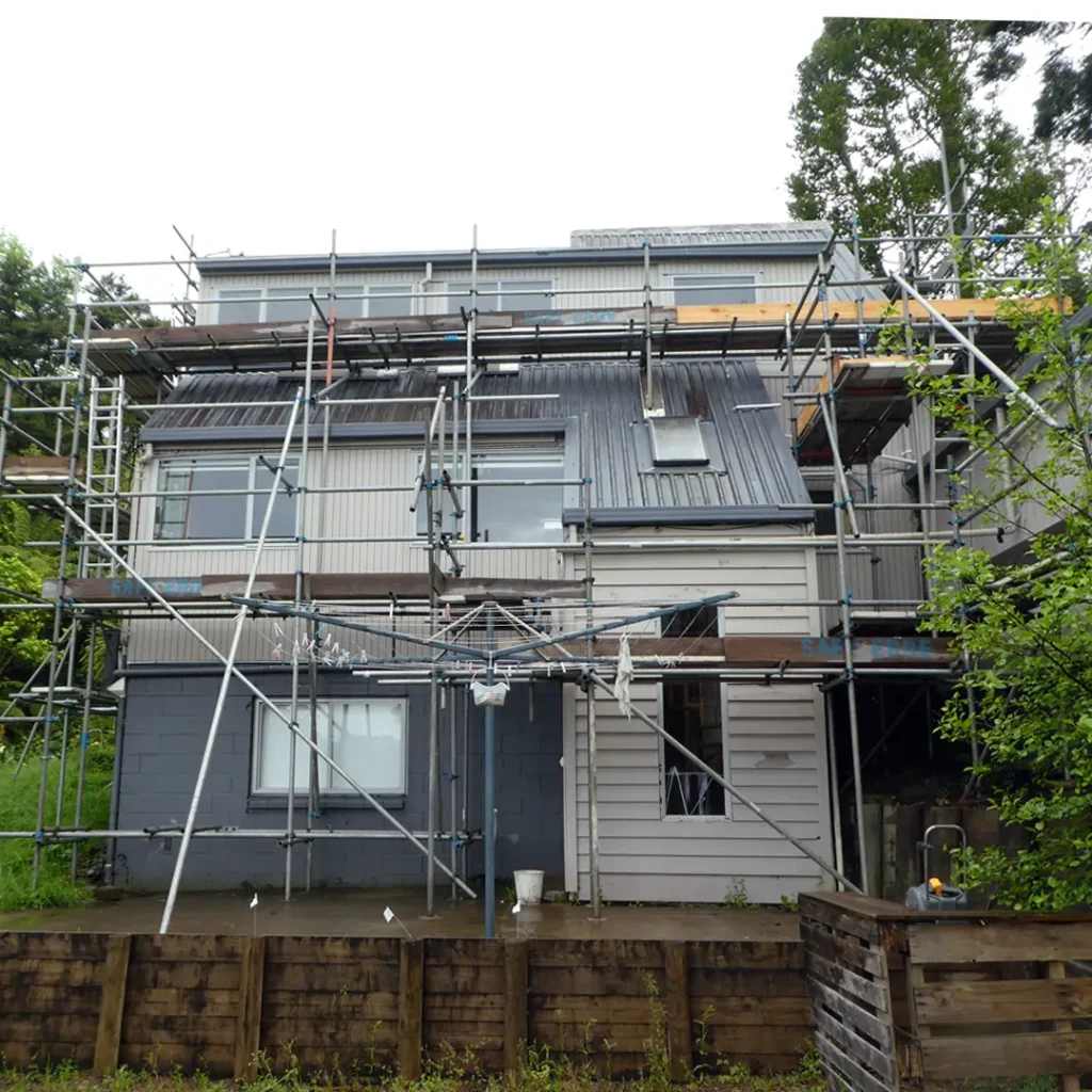 The image is of a house with a retaining wall around it, set against a backdrop of plants, sky, and a tree. The house appears to be undergoing exterior painting as there is scaffolding visible.