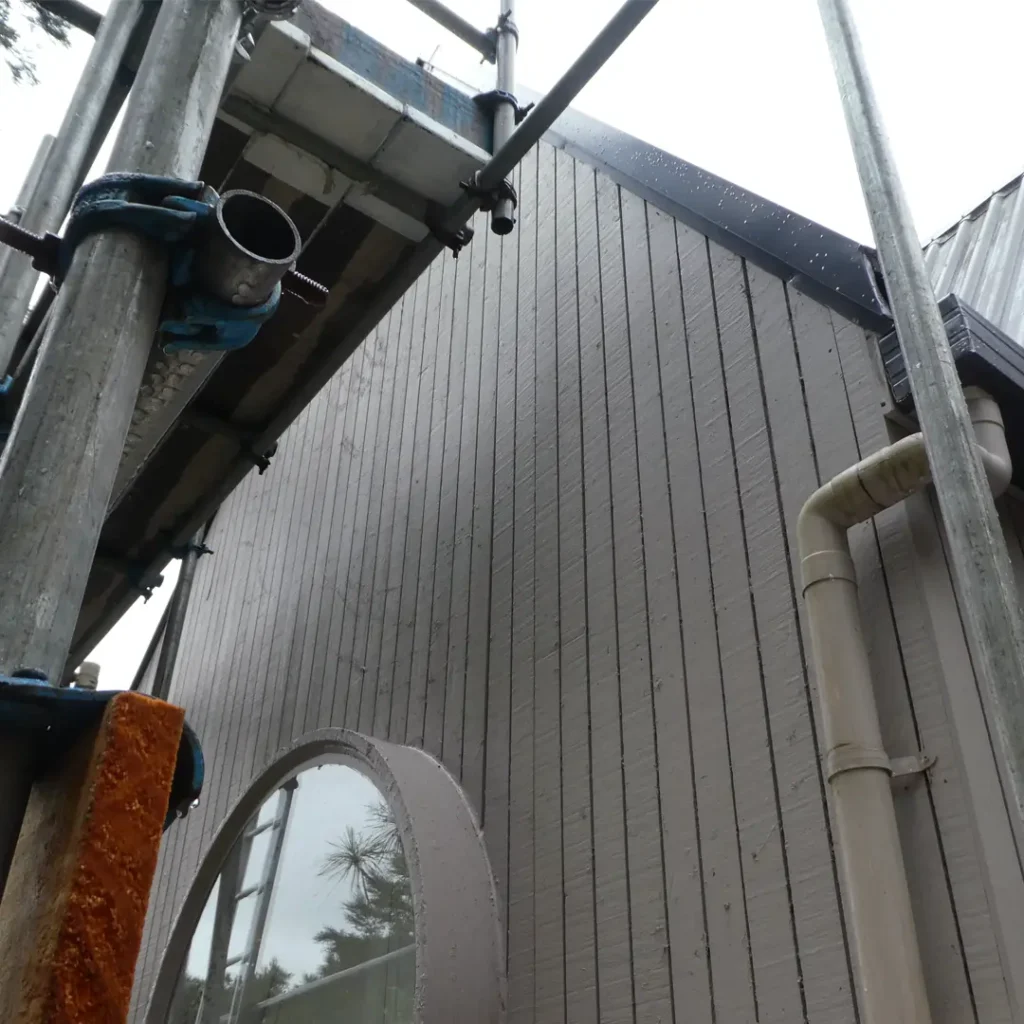 The image depicts a house in preparation for its exterior paint job. The scene is framed by trees and the sky and features a deck, scaffolding, roof, a round window, and vertical cladding.