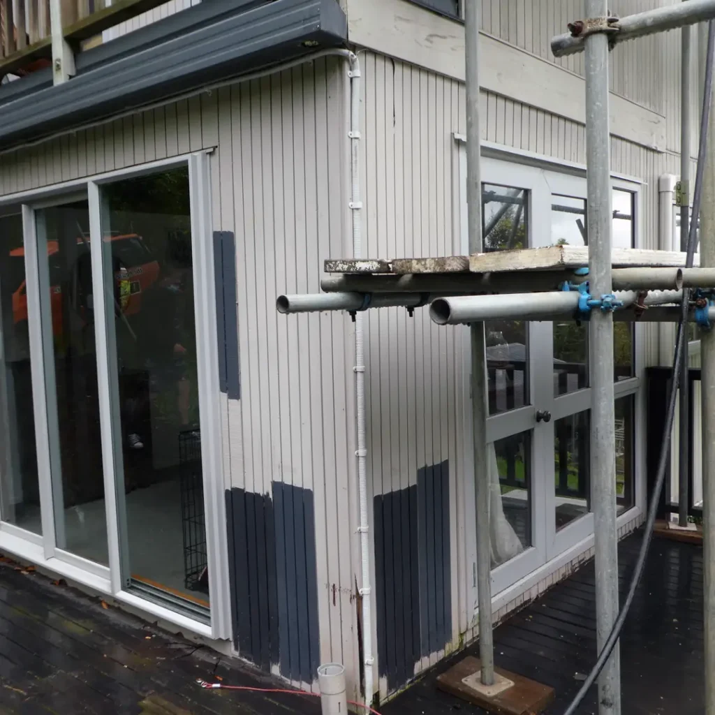 The image depicts a building with scaffolding in front of it. The building appears to be a house with aluminum windows, and it is on the ground level. The context provided suggests that this is before exterior painting. Colour samples have been painted on the walls.