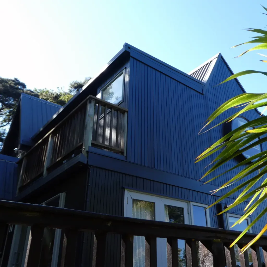 The image depicts a dark house with a tree in front of it, showcasing the exterior after being painted with Weathershield® ColourGuard™ Rawene. It features elements such as aluminium windows, a deck, a round window and vertical cladding.