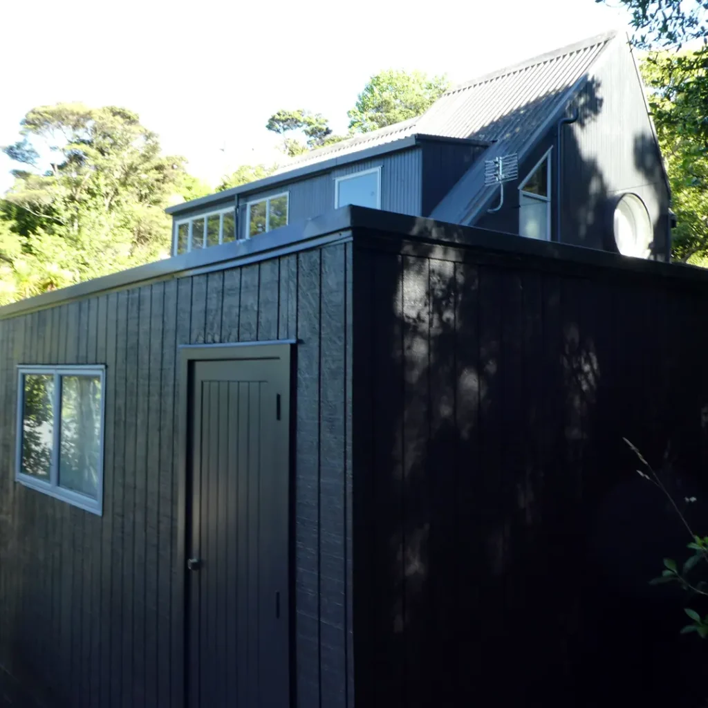 The image shows a house with a door and aluminium windows. It is set outdoors with a blue sky, a tree, and a shed visible. The property has been painted using Weathershield® ColourGuard™ Rawene.