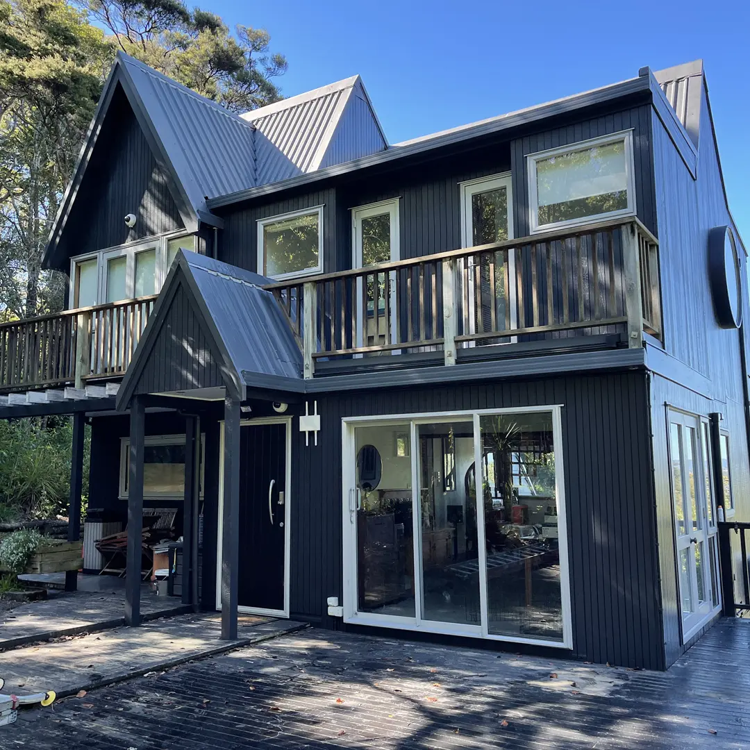 The picture shows the exterior of a darkened house that has been painted with Weathershield® ColourGuardTM Rawene. It has features like vertical cladding, a deck, aluminium windows, and round windows.