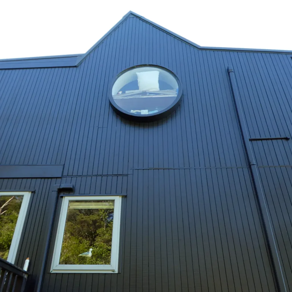 The image depicts a dark house with a tree in front of it, showcasing the exterior after being painted with Weathershield® ColourGuard™ Rawene. It features elements such as aluminium windows, a deck, a round window and vertical cladding.