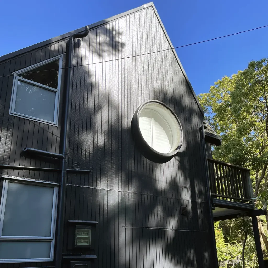 The image depicts a dark house showcasing the exterior after being painted with Weathershield® ColourGuard™ Rawene. It features elements such as aluminium windows, a round window and vertical cladding.