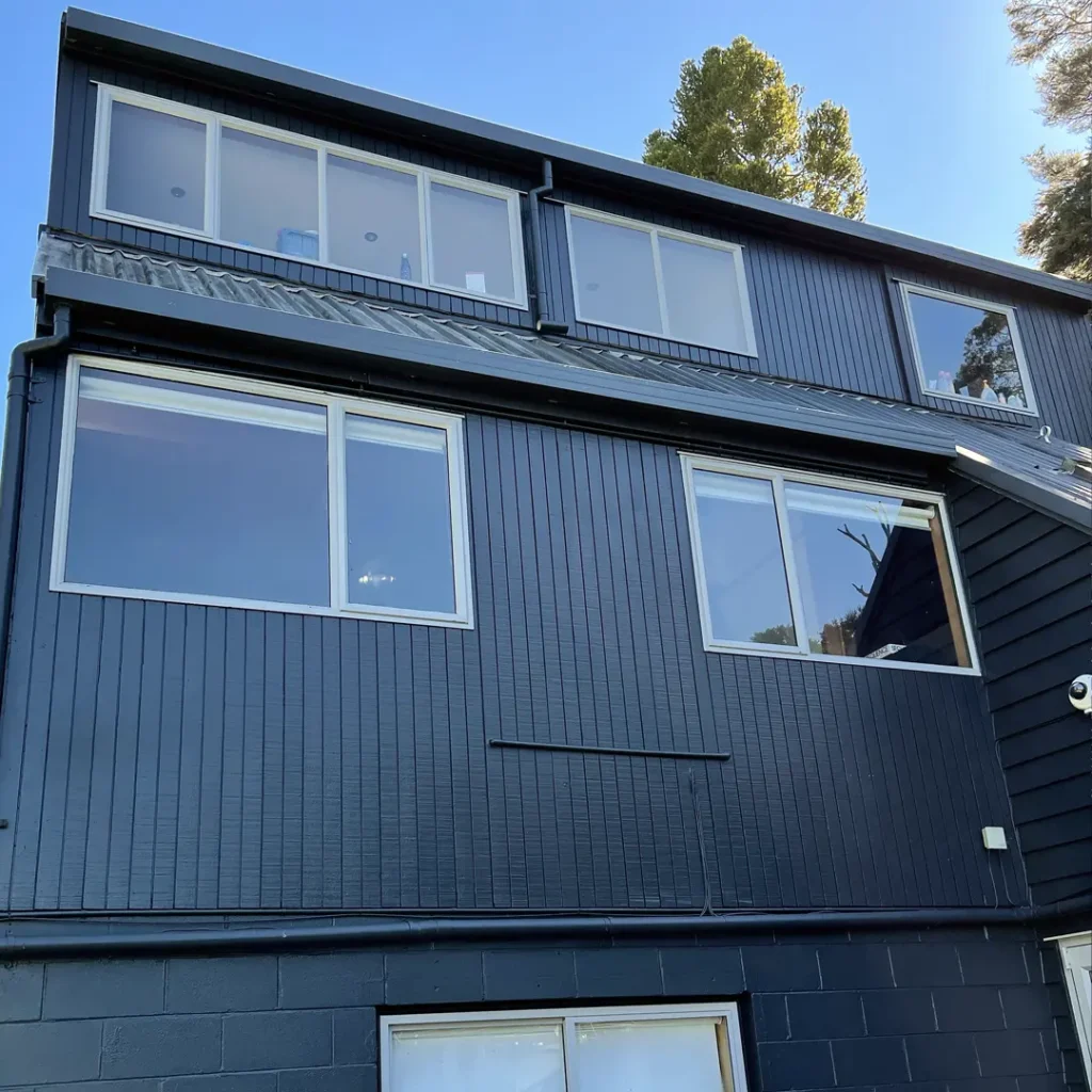 The image depicts a house with aluminium windows, set against a backdrop of the sky, outdoor environment, and a tree. The property appears to have been recently painted using Weathershield® ColourGuard™ Rawene.