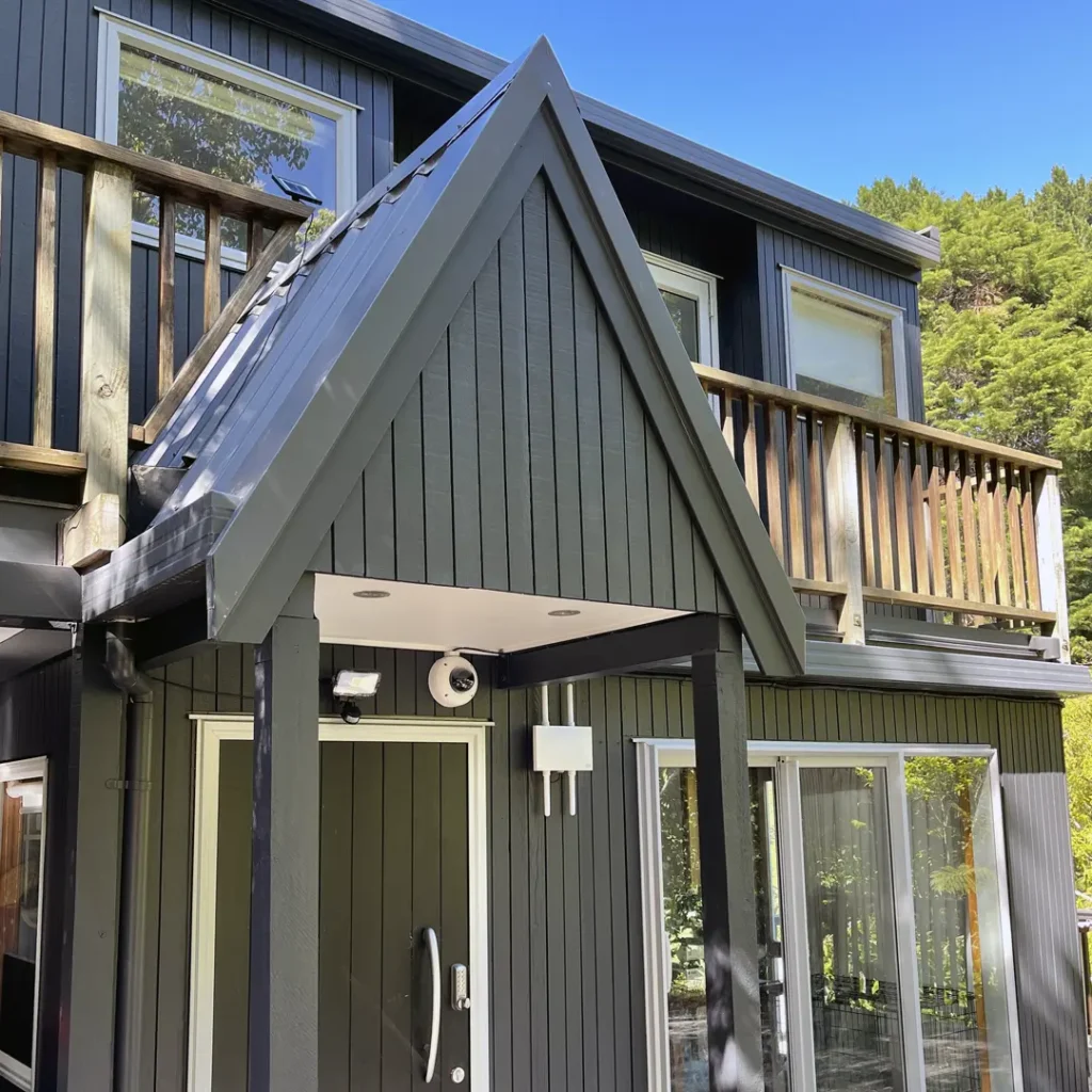 The image depicts a house after exterior painting using Weathershield® ColourGuard™ Rawene. The house features a deck, aluminium windows, vertical cladding, a roof, and is surrounded by trees and a garden.